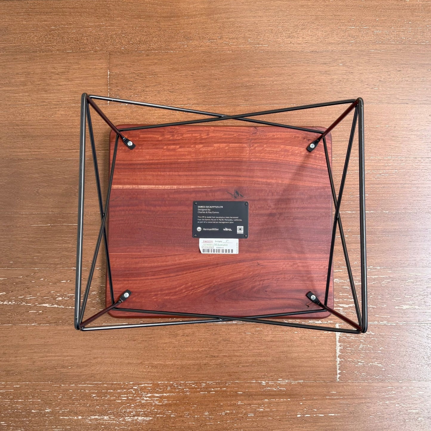 Overhead view of the Eames Eucalyptus LTR Table showing its solid eucalyptus wood top with natural knots and cracks, supported by a black powder-coated wire base on a wooden floor.