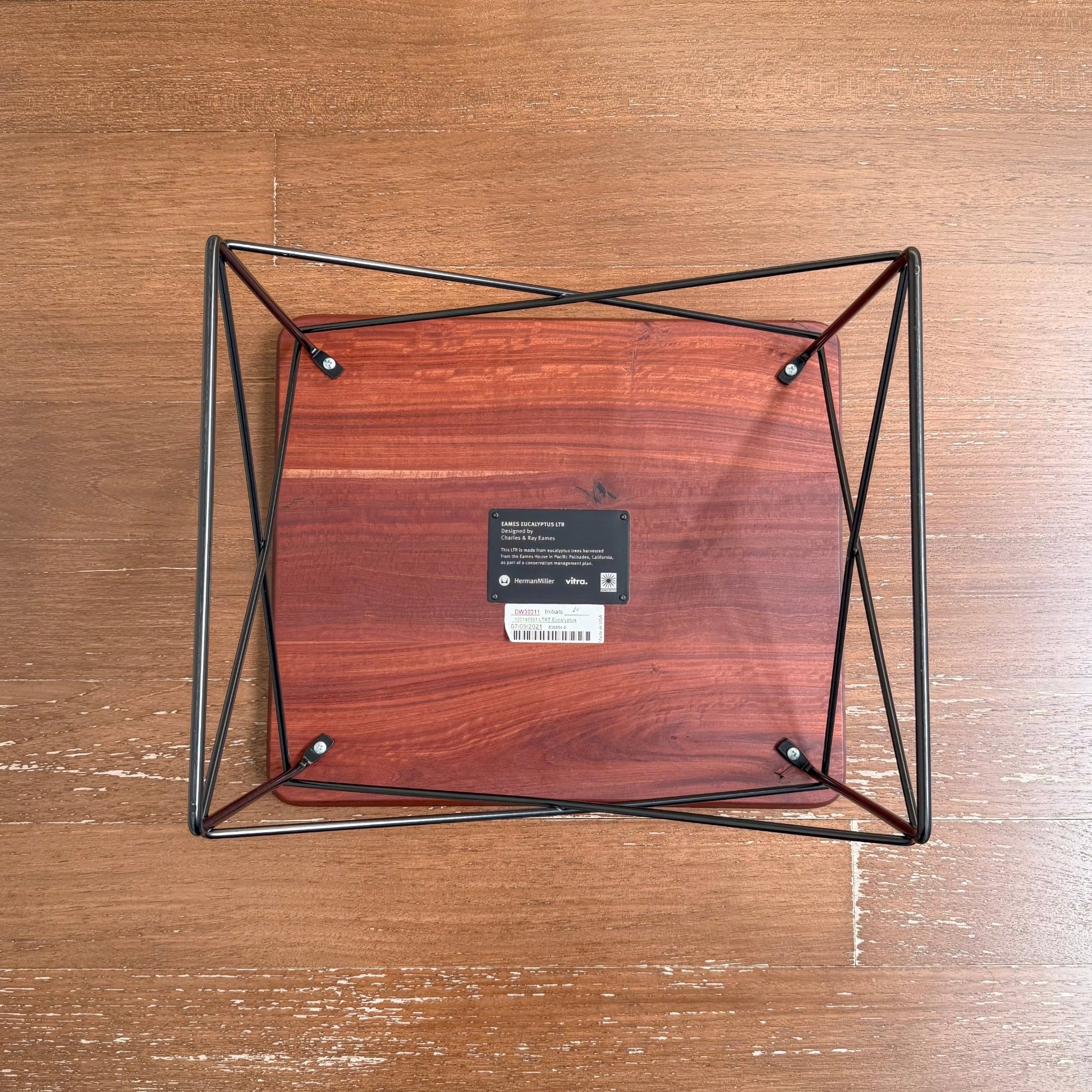 Overhead view of the Eames Eucalyptus LTR Table showing its solid eucalyptus wood top with natural knots and cracks, supported by a black powder-coated wire base on a wooden floor.