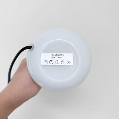 Hand holding the base of a Holmegaard Carol White Opal Glass Table Lamp, showing the underside label and attached black cord against a plain background.