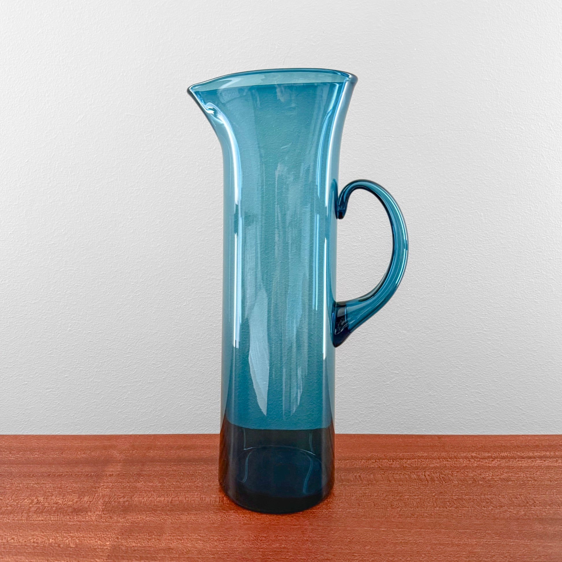 Ingeborg Lundin Orrefors Blue Glass Pitcher standing on a wooden surface, featuring a tall, slender design with a curved handle and a deep blue translucent finish.