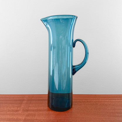Ingeborg Lundin Orrefors Blue Glass Pitcher standing on a wooden surface, featuring a tall, slender design with a curved handle and a deep blue translucent finish.