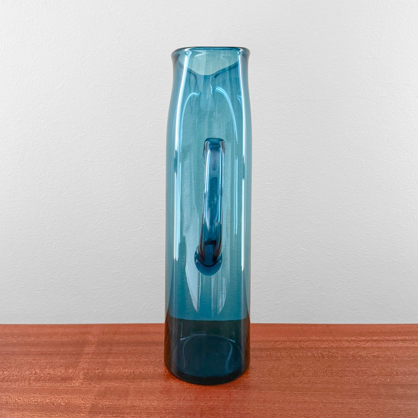 Ingeborg Lundin Orrefors Blue Glass Pitcher standing on a wooden surface, showcasing its tall, slender cylindrical shape with a smooth handle, crafted in vintage Scandinavian glass design.
