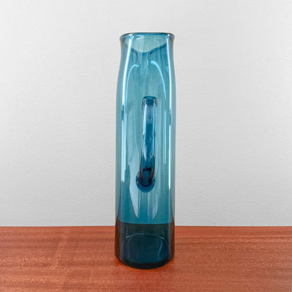 Ingeborg Lundin Orrefors Blue Glass Pitcher standing on a wooden surface, showcasing its tall, slender cylindrical shape with a smooth handle, crafted in vintage Scandinavian glass design.