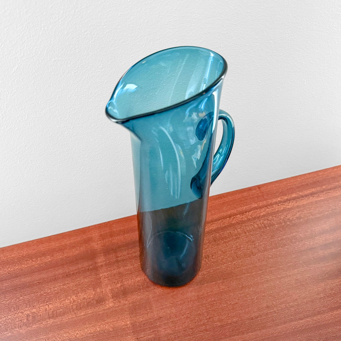 Ingeborg Lundin Orrefors Blue Glass Pitcher standing on a wooden surface, showcasing its sleek vintage Scandinavian design and curved handle.