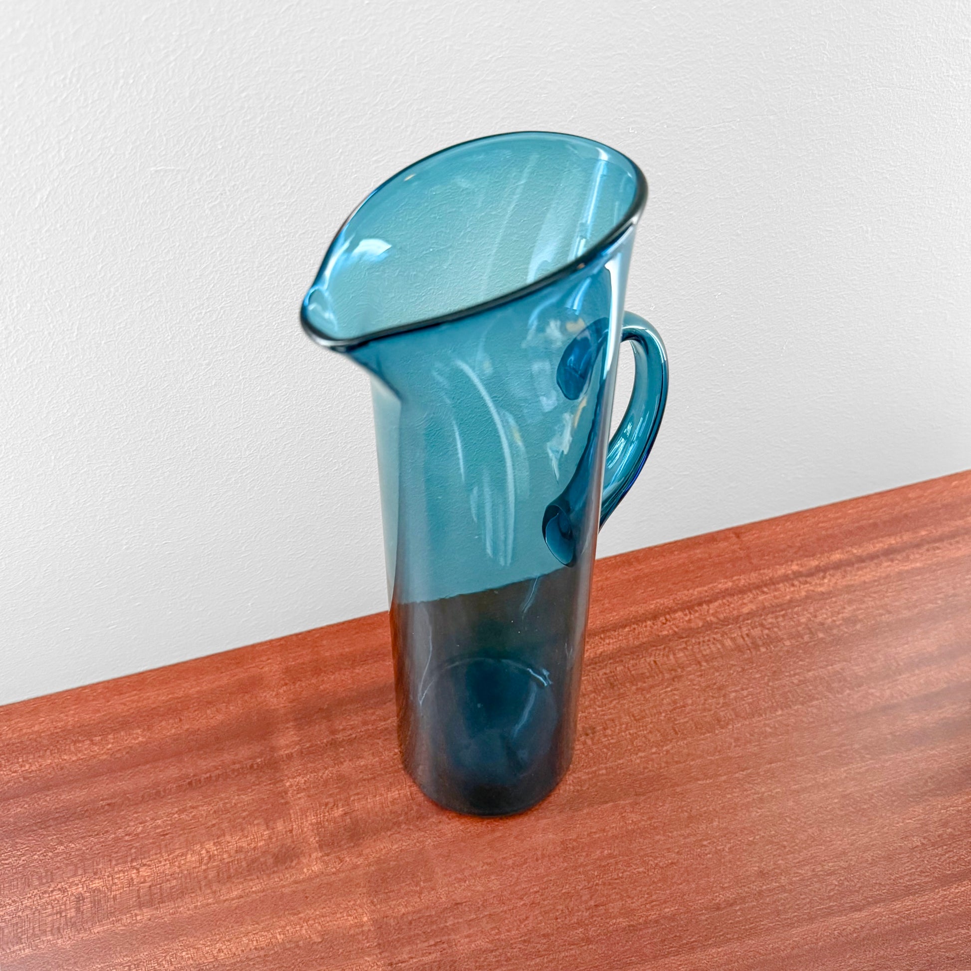 Ingeborg Lundin Orrefors Blue Glass Pitcher standing on a wooden surface, showcasing its sleek vintage Scandinavian design and curved handle.
