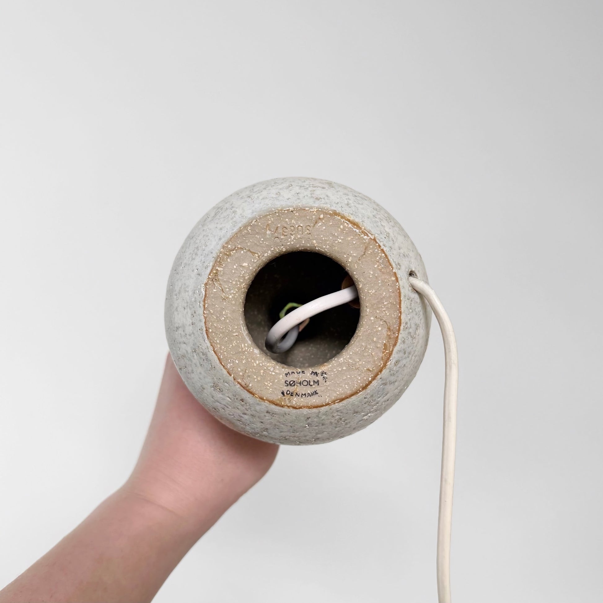 Hand holding the underside of a Søholm Erika Small Stoneware Table Lamp showing the ceramic base with maker's stamp and electrical cord visible against a white background.
