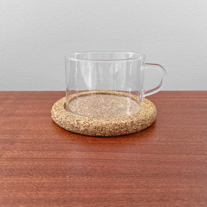Signe Persson-Melin Boda Nova Glass Tea Cup with Cork Coaster featuring a clear glass cup with handle resting on a round natural cork coaster, displayed on a wooden surface.