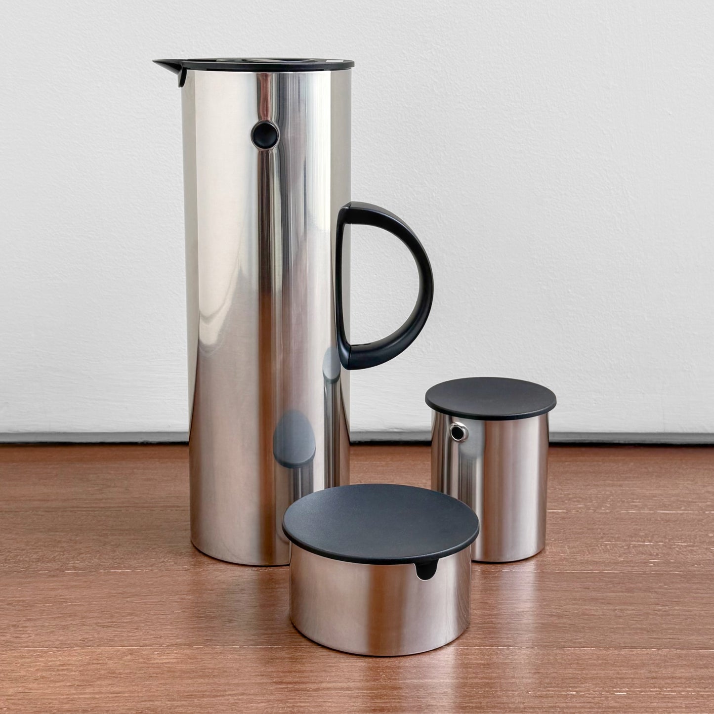 Stelton EM77 Stainless Steel Coffee Set featuring a tall vacuum jug with black handle and lid, accompanied by a sugar bowl and creamer with black lids, displayed on a wooden surface.